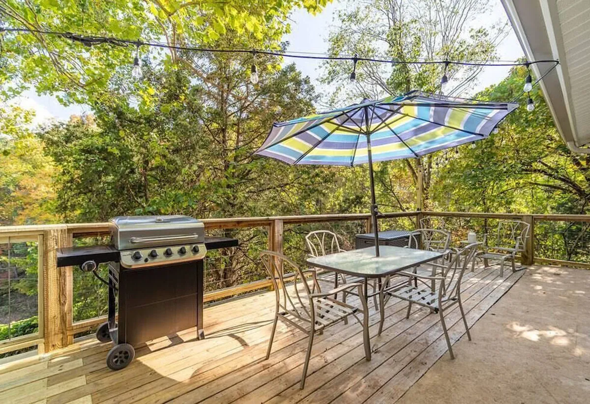 An exterior shot of a spacious wooden deck featuring a grill, dining table with umbrella, and ample seating. The deck overlooks natural surroundings, offering an inviting spot for outdoor dining, relaxing, and social gatherings, especially during sunset.