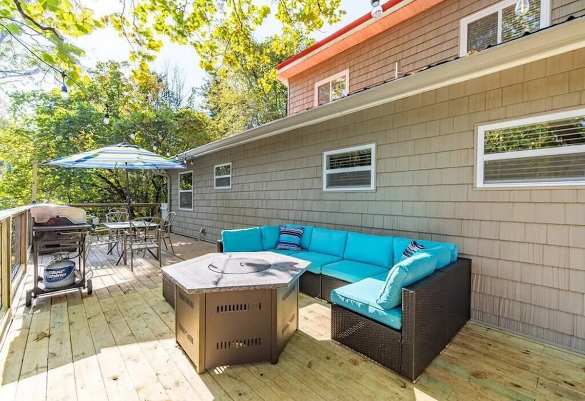 An inviting outdoor living area featuring a fire pit encircled by comfortable seating, a grill for barbecues, and a large umbrella providing shade. The deck area is designed for socializing, sharing meals, and enjoying sunset views with friends or extended family.