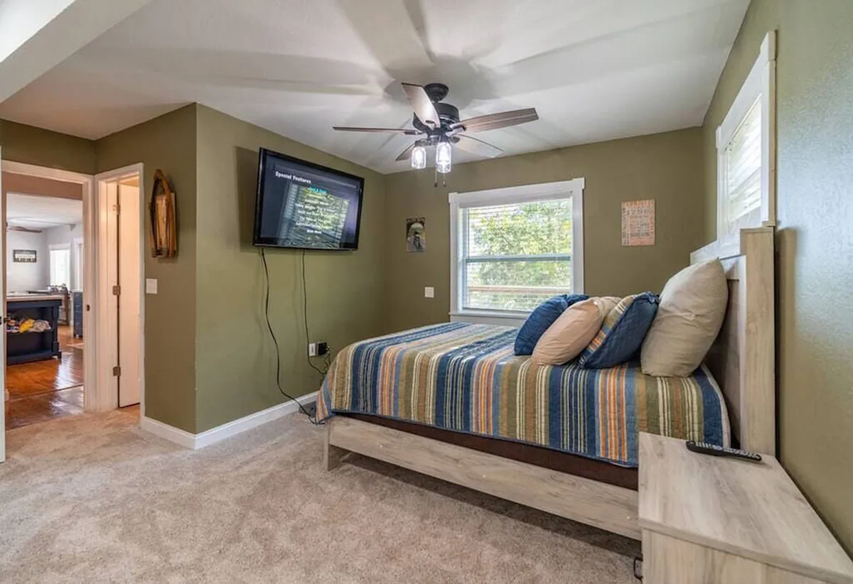 A cozy bedroom with striped bedding, a wall-mounted Smart TV, and a large window inviting in natural light. The layout suggests a welcoming space for restful sleep, movie nights, or unwinding after a lakeside stroll. The window connects the interior to the outside world, maintaining the lakeside retreat feel.