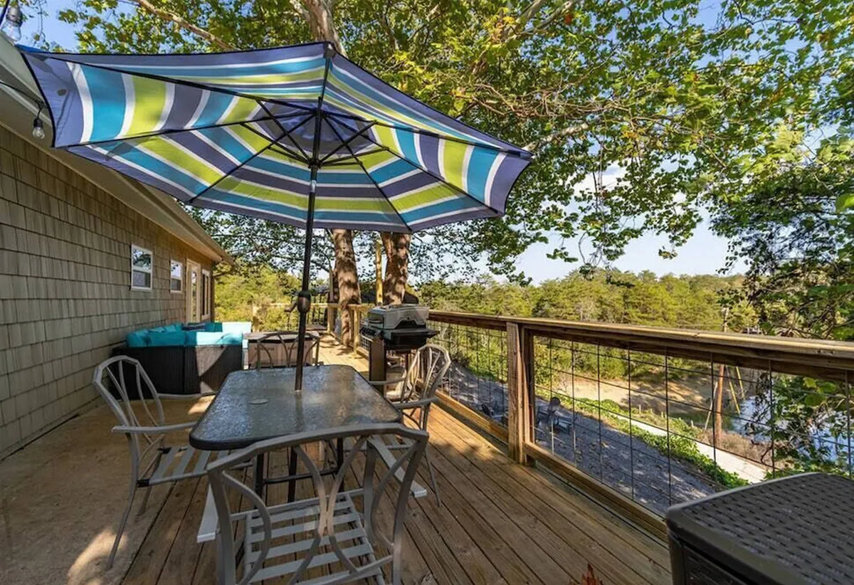 This exterior shot features a colorful umbrella shading an outdoor dining table, with lush trees creating a tranquil, natural atmosphere. The scene radiates a sense of relaxation and connection to the outdoors—perfect for couples or small groups enjoying a meal or a quiet morning coffee together. The grill in the background hints at the possibility of leisurely barbecues and shared experiences in a private, scenic setting.