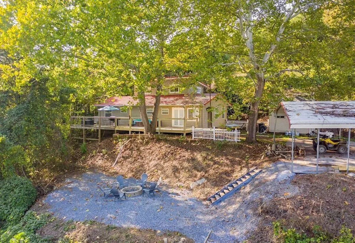 An inviting exterior view of a house with a porch, a cozy fire pit area, and trees surrounding the property. The scene suggests communal outdoor activities like gathering around the fire and enjoying the natural surroundings.