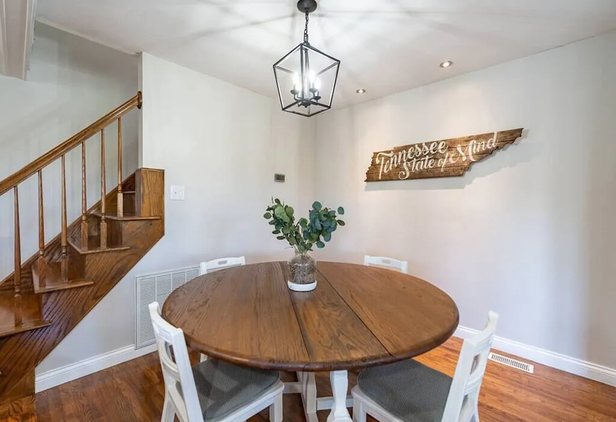 A cozy dining area with a round wooden dining table, a green plant centerpiece, and a decorative wall sign near a wooden staircase. The scene is set for intimate family meals, lively conversations, or festive celebrations, radiating a welcoming atmosphere.