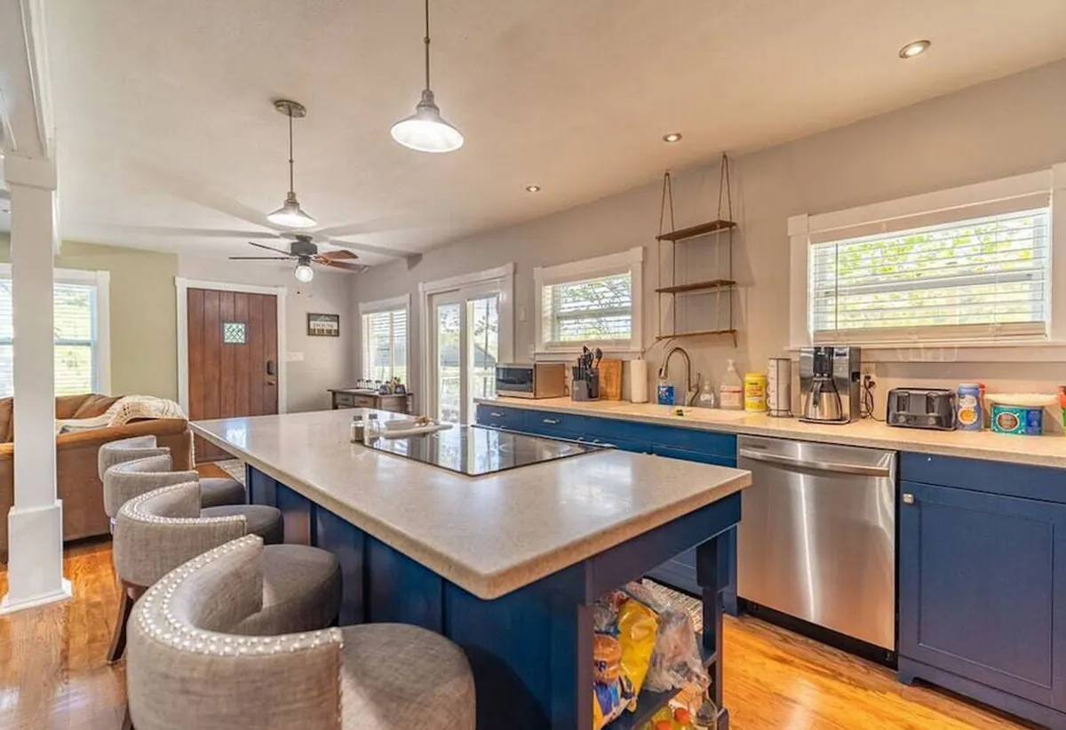 This image showcases a warm, welcoming kitchen featuring an island countertop, blue cabinets, bar stools, and modern kitchen appliances. The setup invites both cooking and socializing, with room for group activities around the island and plenty of prep space.