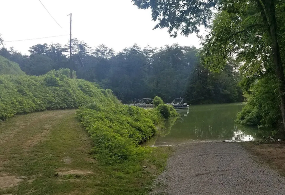 A tranquil lakeside scene featuring a gravel path leading through lush greenery to the calm water’s edge. The serene setting suggests opportunities for boating, fishing, walking, and picnicking, with subtle hints of wildlife and sunset reflections over the water.