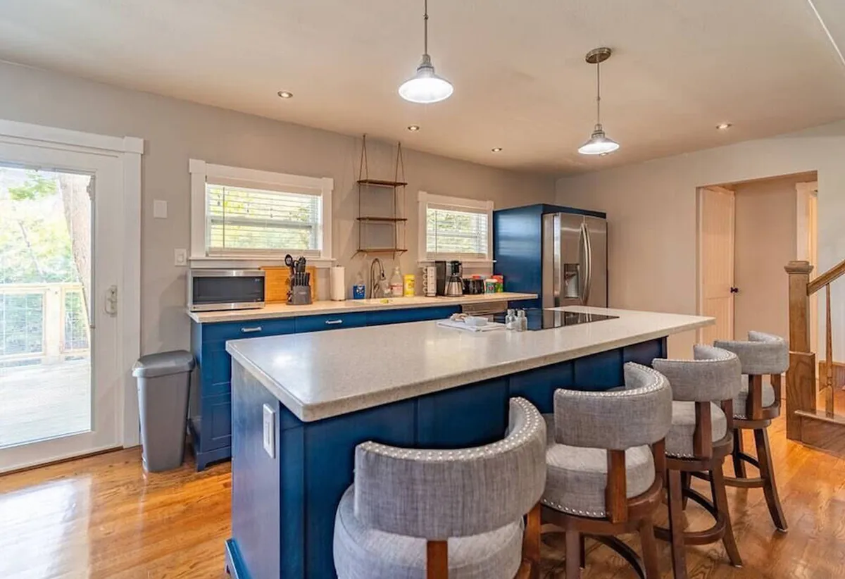 A bright, welcoming kitchen scene featuring a large kitchen island with bar stools, ample countertop space, modern appliances, and plenty of windows for natural light. The setup encourages both cooking and casual dining, with space for multiple people to gather and participate.