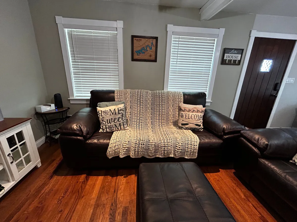 A close-up of a leather sofa adorned with decorative pillows and a woven blanket, accompanied by a wooden coffee table and a doorway to the balcony. The space is designed for relaxing, reading, socializing, and watching TV, exuding an inviting mood for both quiet moments and gatherings.