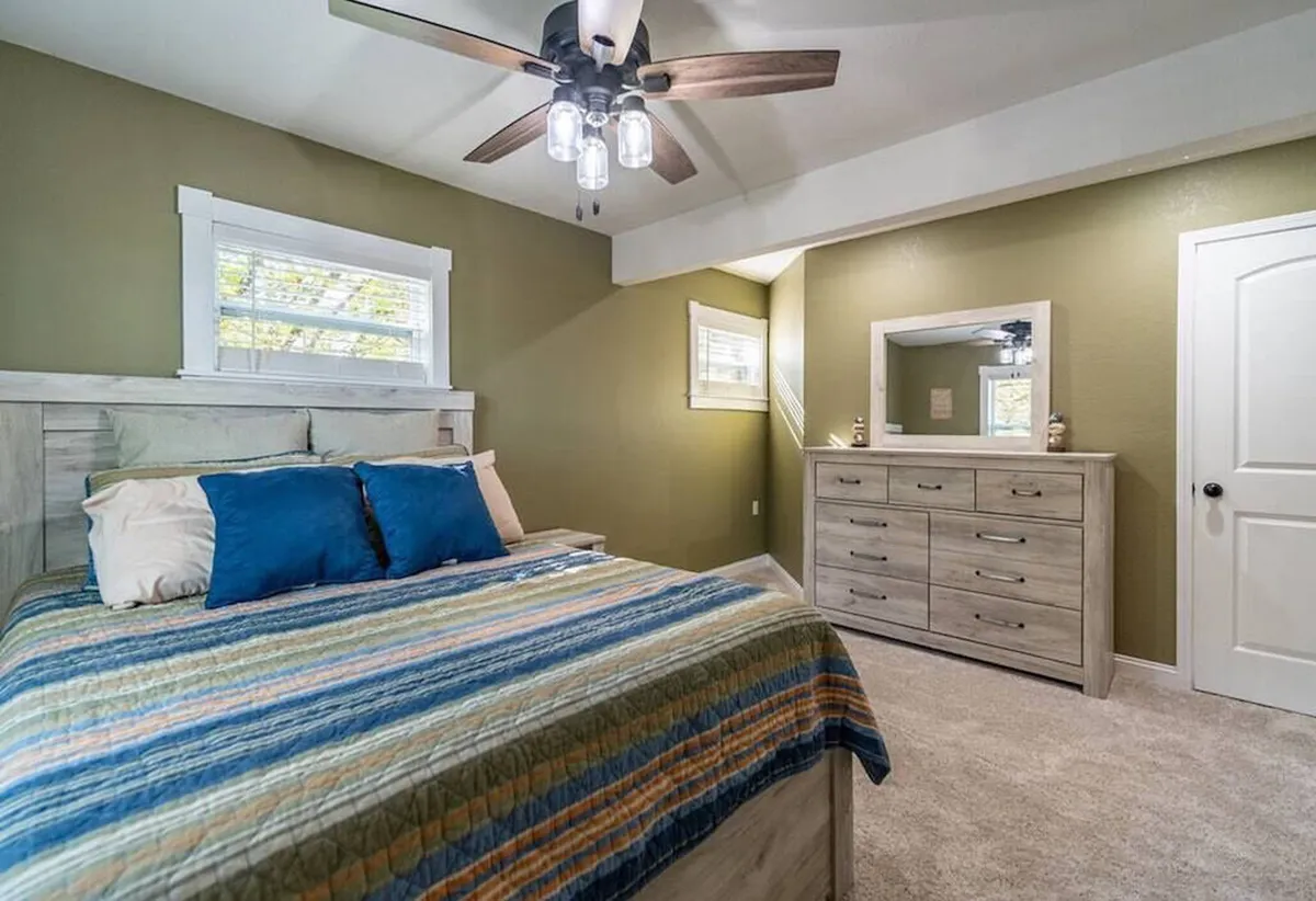 A tranquil bedroom featuring a bed with colorful bedding, a dresser with mirror, and a ceiling fan, bathed in natural light from the window. The room exudes a cozy, inviting atmosphere, perfect for restful nights and quiet mornings.